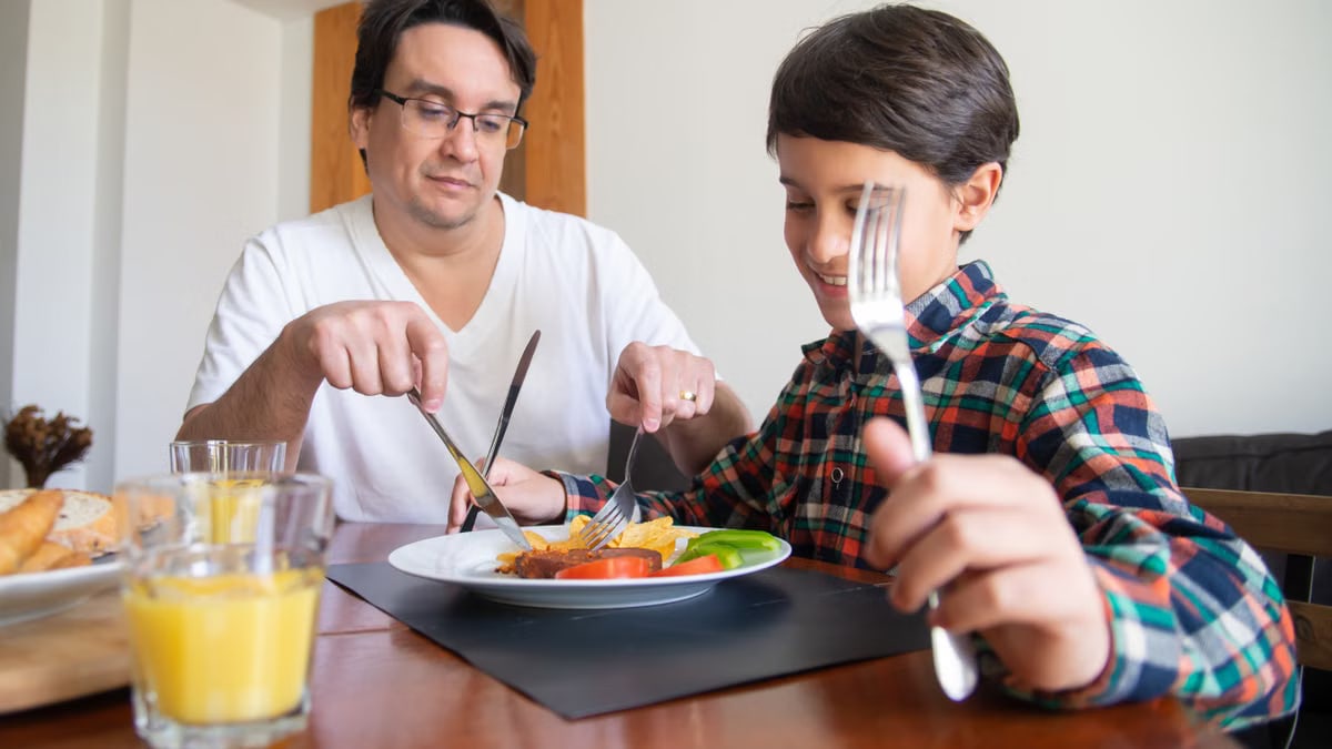 A father and son share a meal indoors, practicing dining etiquette with orange juice and healthy options.